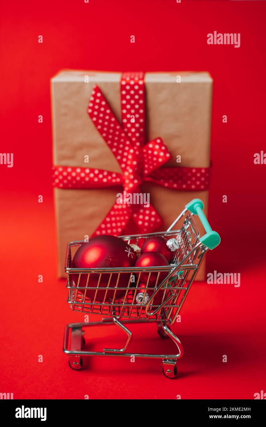 Vente de Noël bannière Web avec mini chariot avec boîte cadeau et décorations de Noël rouges boules sur fond rouge. Achats de Noël minimaux Banque D'Images