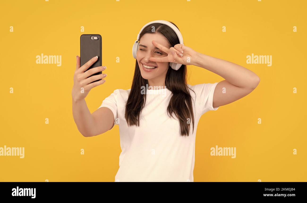 femme positive en chemise blanche dans le casque faisant selfie sur le téléphone avec le geste de paix, selfie Banque D'Images