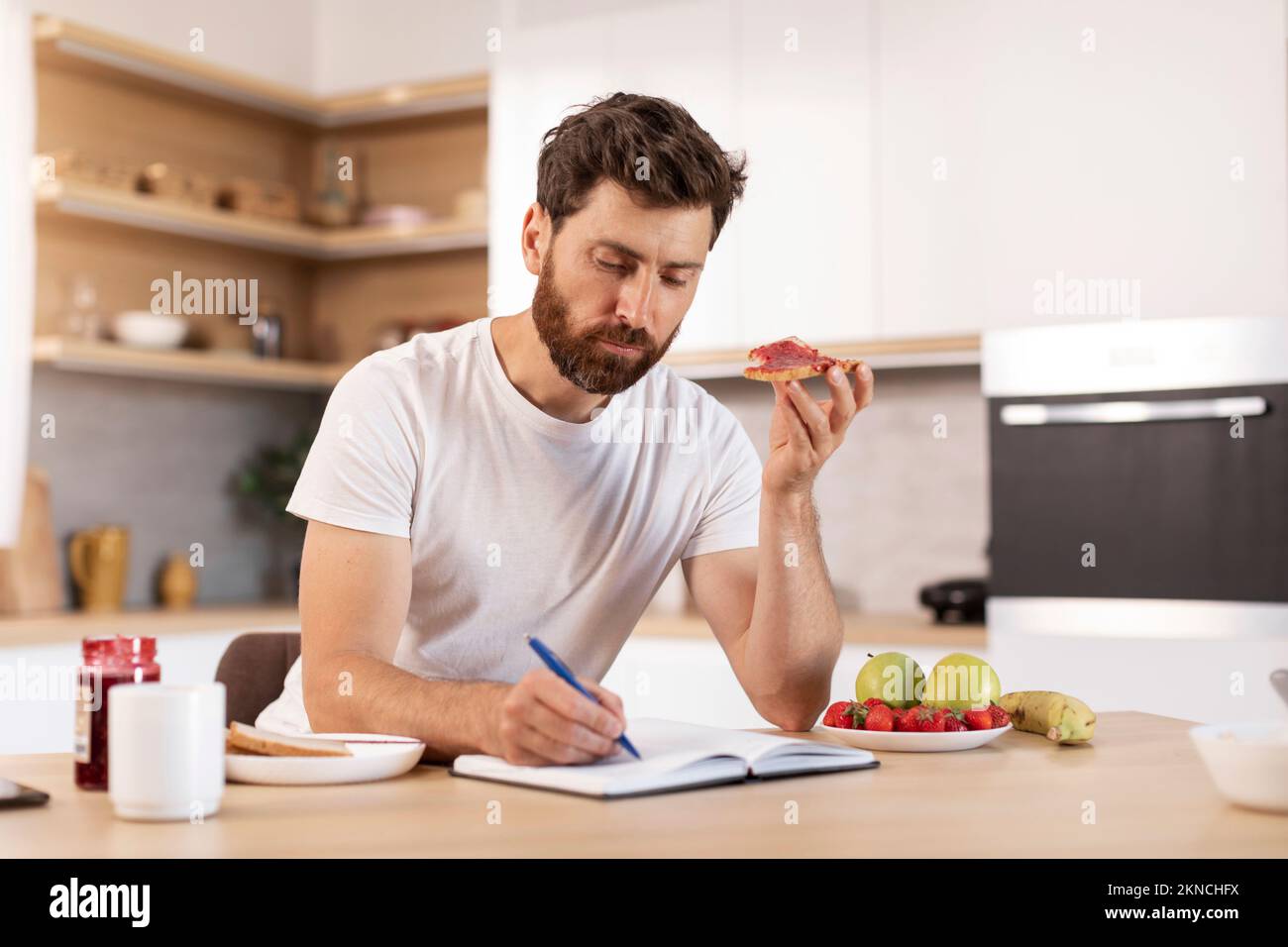 Homme caucasien adulte concentré occupé avec une barbe en t-shirt blanc mange un sandwich, fait des notes dans le carnet Banque D'Images