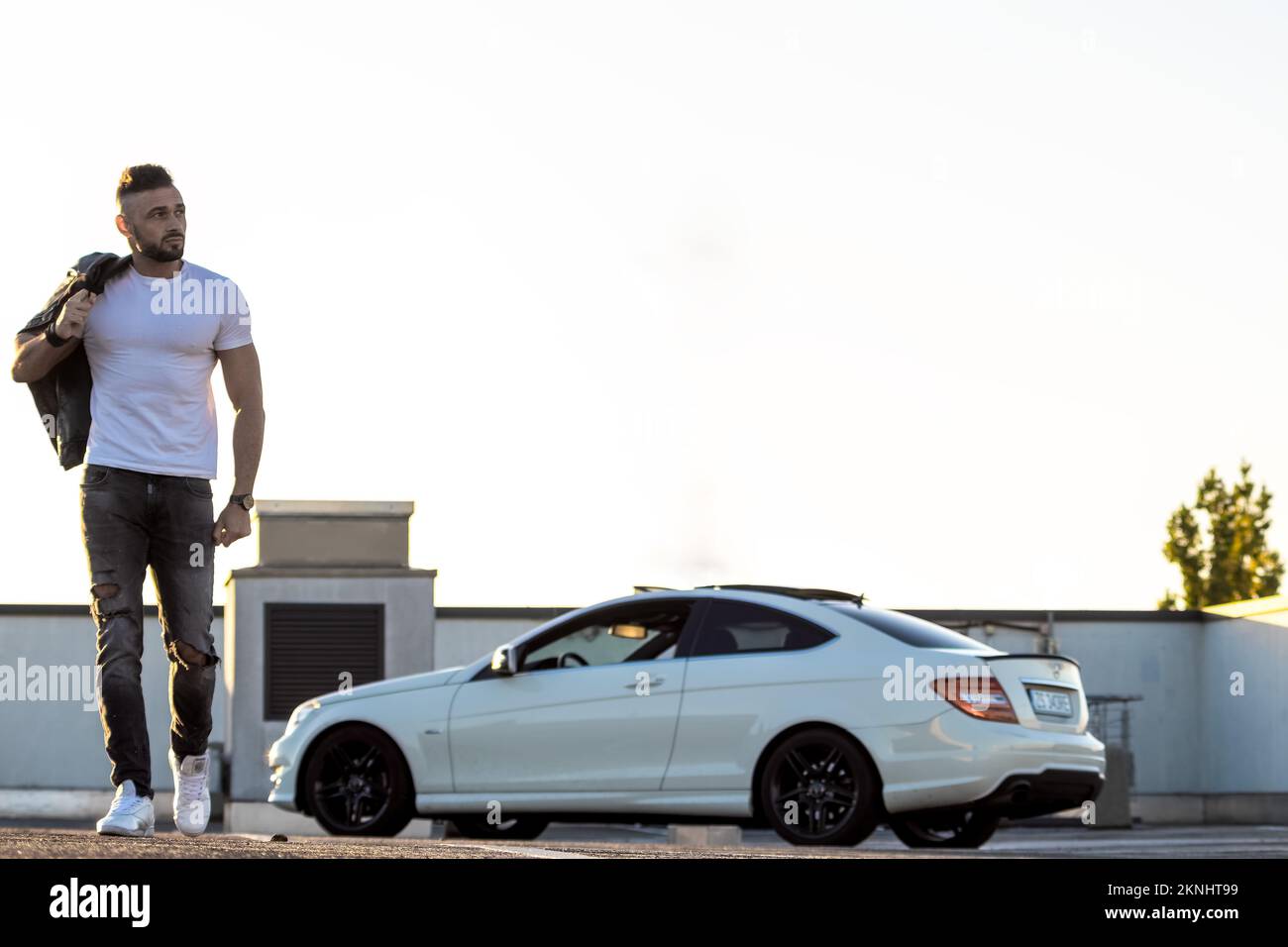 Un homme dur dans un T-shirt blanc s'éloigne d'une voiture blanche et regarde loin. Banque D'Images