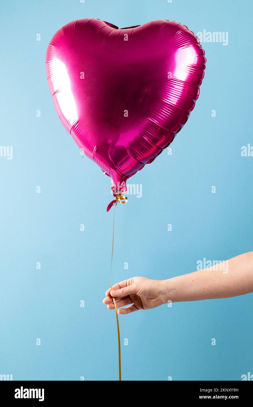 Image verticale de la main tenant un ballon rose brillant en forme de coeur, flottant sur fond bleu avec espace de copie Banque D'Images