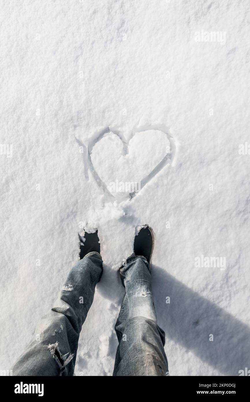 Détails verticaux sur les jambes d'un homme voyageur debout dans le point de vue de la 1ère personne avec coeur tiré dans la neige. Les pieds froids ou l'amour des vacances d'hiver Banque D'Images
