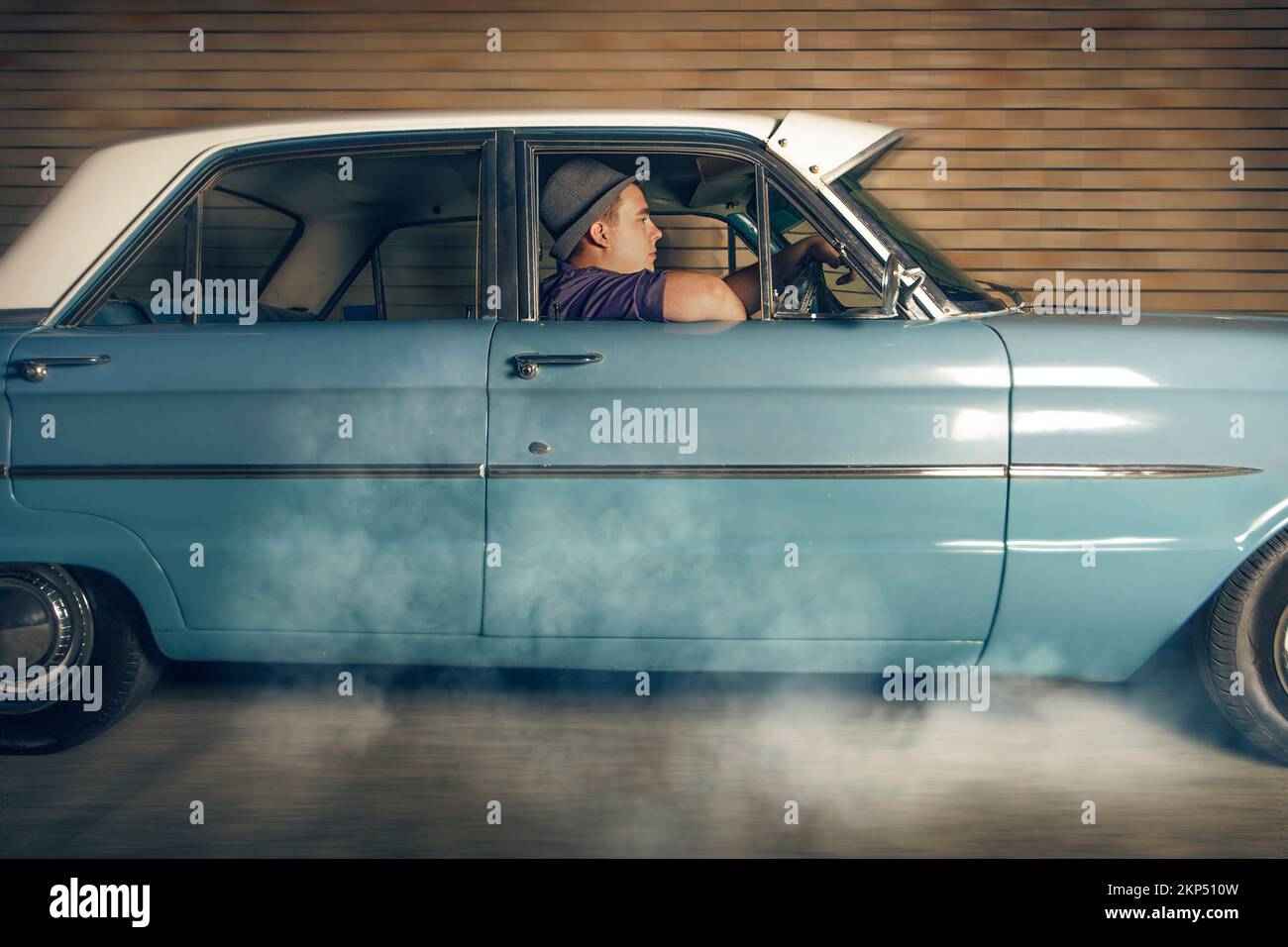 Un homme de 1950 ans assis à l'intérieur d'une voiture d'époque fume les pneus dans un mouvement de décollage en s'éloignant du passage de la voie arrière. Poursuite à chaud Banque D'Images