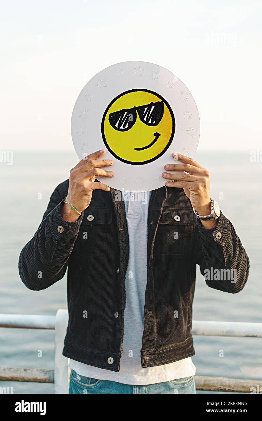 Un jeune homme adulte couvre le visage avec un visage emoji souriant avec des lunettes de soleil, dessiné à la main sur un disque blanc debout à l'extérieur contre le réseau social de la mer dans Banque D'Images