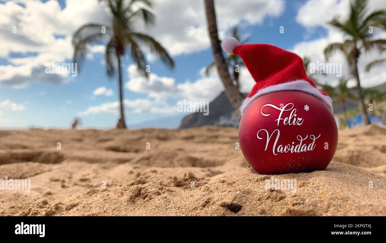 Bombe de Noël dans le chapeau du Père Noël avec des mots Joyeux Noël en espagnol sur la plage sur le sable avec des palmiers et le ciel bleu sur le fond. Banque D'Images