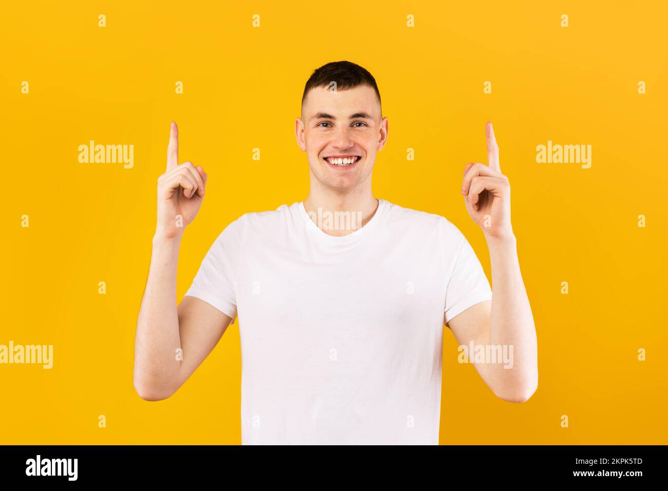 Joyeux caucasien dans un t-shirt blanc pointant vers le haut de l'espace vide et souriant à la caméra, debout sur fond jaune Banque D'Images