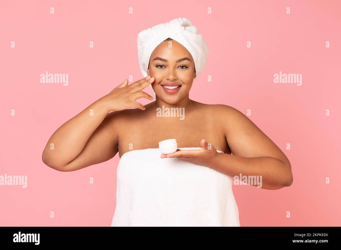 Concept de soin quotidien de la peau. Portrait d'une femme afro-américaine en surpoids tenant un pot de crème et appliquant un hydratant sur la joue Banque D'Images