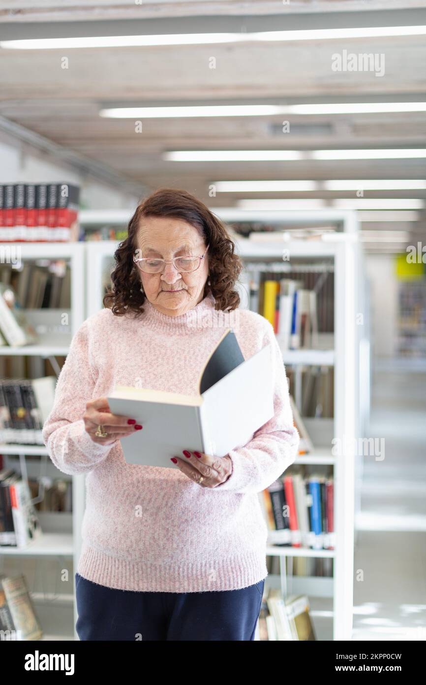 Femme caucasienne âgée lisant un livre dans une bibliothèque publique. Concept de divertissement des personnes âgées. Banque D'Images