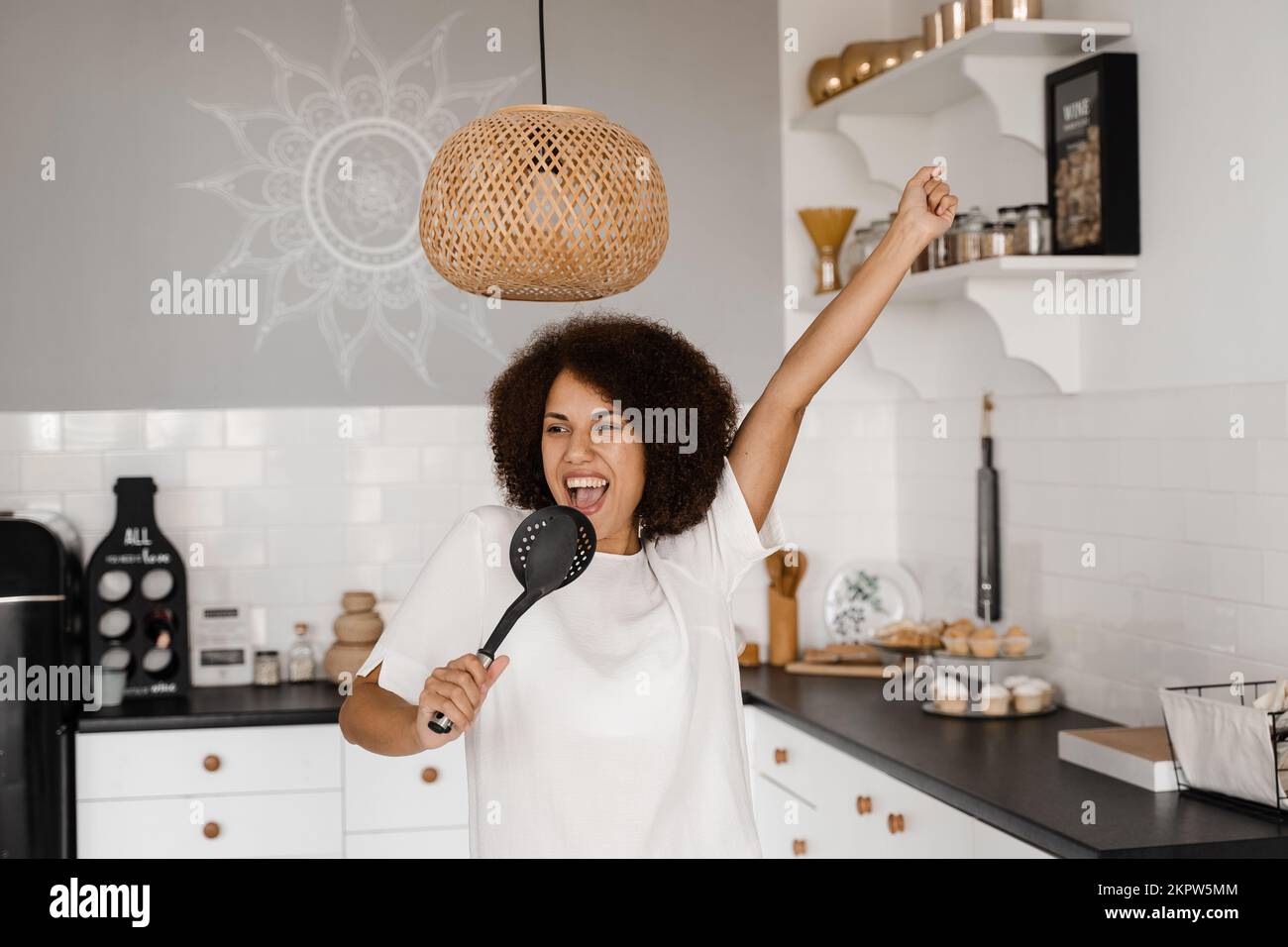 Femme de ménage afro-américaine en tablier chantant avec des spatules pour la cuisson du microphone sur la cuisine. Joyeuse jeune femme africaine s'amusant avec cuisine ut Banque D'Images
