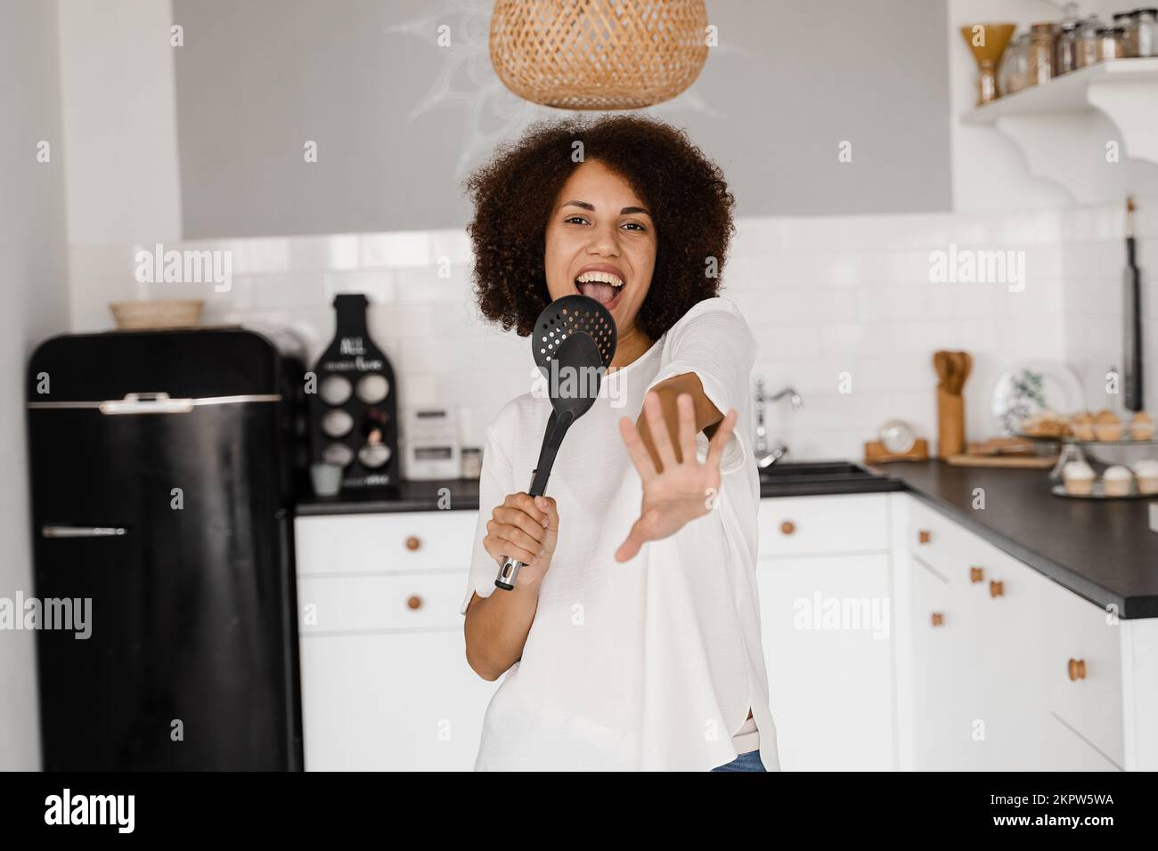 Joyeuse jeune femme africaine s'amusant avec des ustensiles de cuisine. Femme de ménage afro-américaine en tablier chantant avec des spatules pour la cuisson du microphone sur le k Banque D'Images