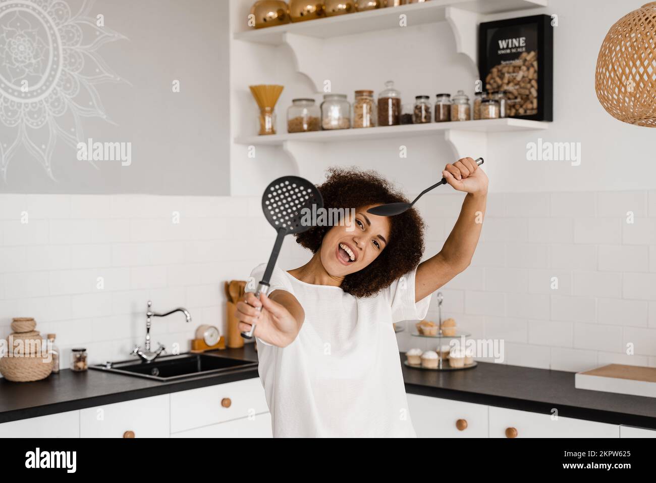 La femme de ménage africaine en tablier pose avec des spatules pour cuisiner sur la cuisine. Afro-américaine fille cuisinière s'amuser dans la cuisine. Publicité pour c Banque D'Images