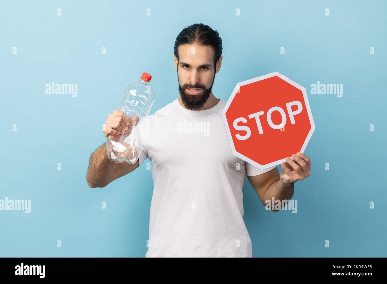 Portrait d'un homme responsable avec une barbe portant un T-shirt blanc tenant une bouteille en plastique et un panneau rouge stop, se préoccupant de l'environnement. Studio d'intérieur isolé sur fond bleu. Banque D'Images