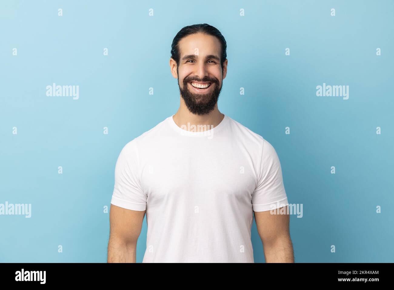 Portrait d'un beau homme barbu portant un T-shirt blanc debout regardant l'appareil photo avec un visage satisfait et souriant, exprimant le bonheur. Studio d'intérieur isolé sur fond bleu. Banque D'Images