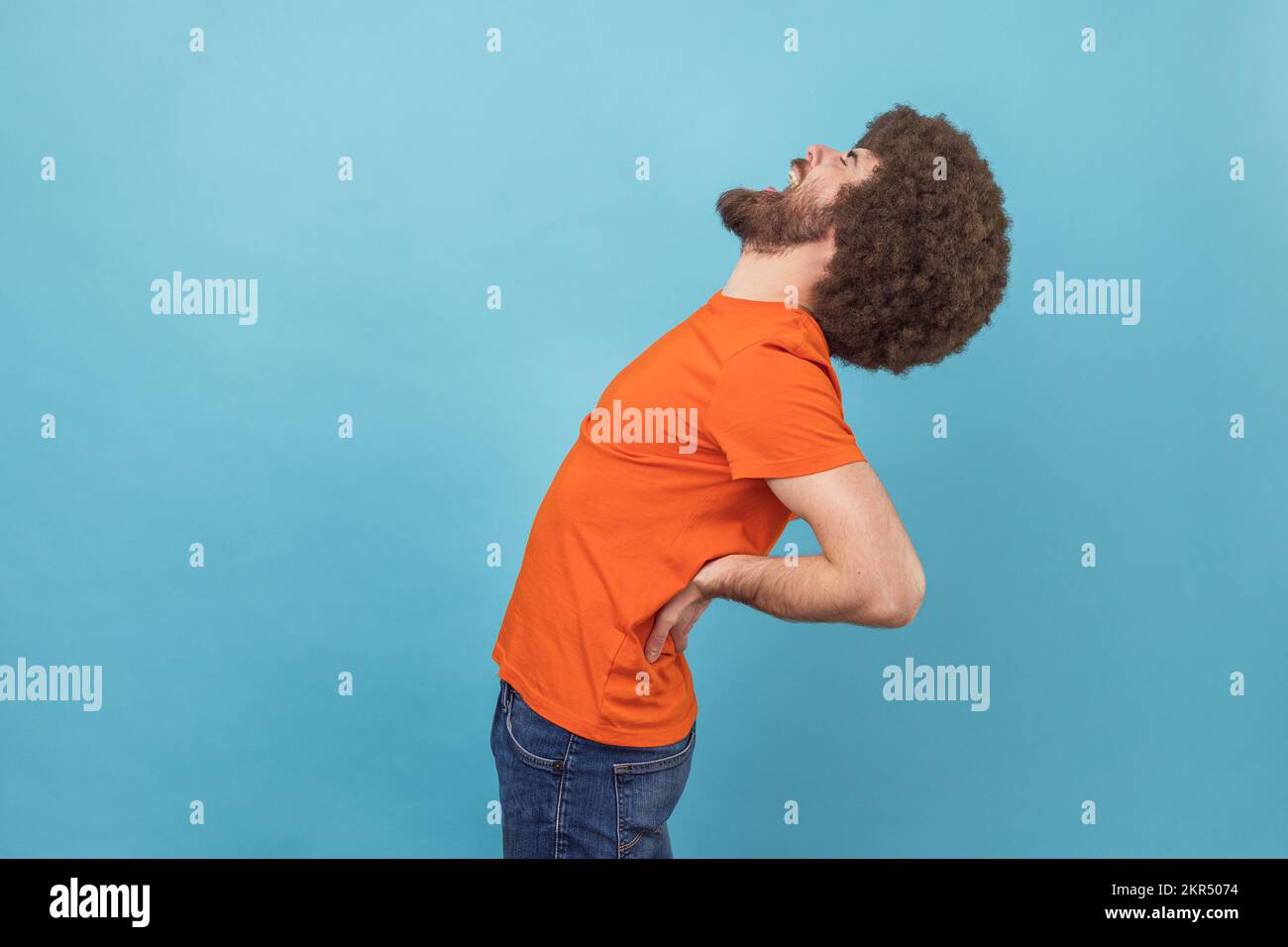 Vue latérale portrait d'un homme avec une coiffure afro portant un T-shirt orange debout avec des yeux fermés, souffrant de douleurs dans le bas du dos, une inflammation des reins. Studio d'intérieur isolé sur fond bleu. Banque D'Images