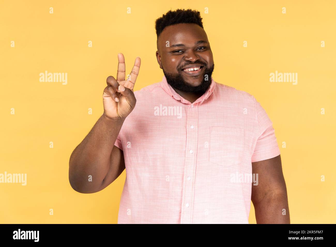 Portrait de l'homme portant une chemise rose faisant un geste de victoire et regardant playfully à l'appareil photo, montrant la paix, le signe v avec des doigts doubles. Studio d'intérieur isolé sur fond jaune. Banque D'Images