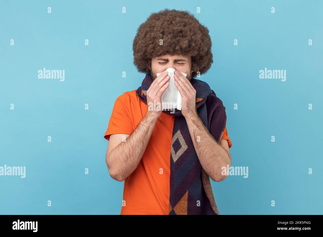 Portrait d'un homme malsain avec une coiffure afro portant un T-shirt orange debout, tenant le tissu sur la bouche et le nez, enveloppé dans une écharpe chaude. Studio d'intérieur isolé sur fond bleu. Banque D'Images