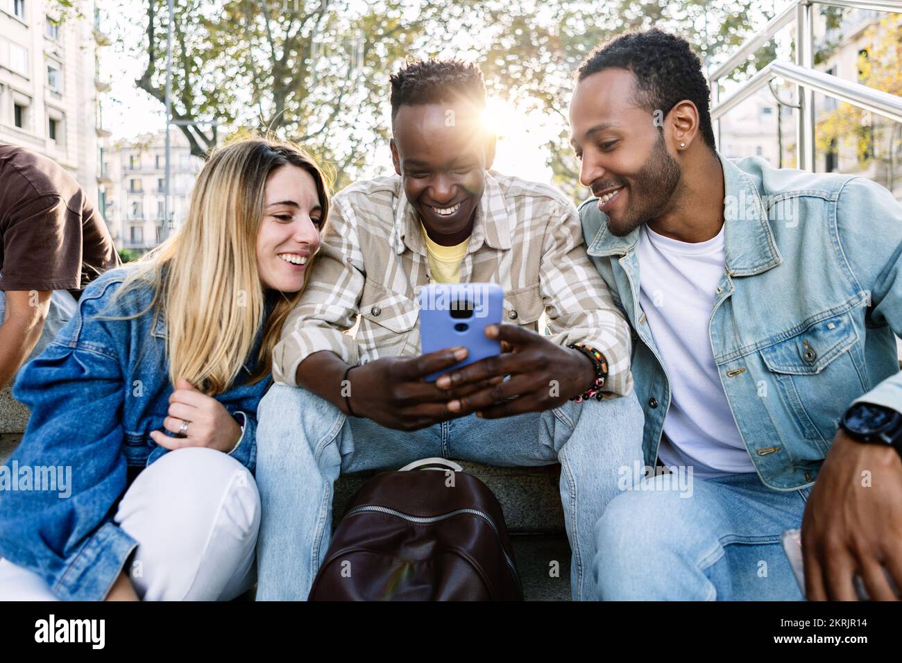 Groupe de jeunes amis multiethniques utilisant un téléphone portable assis ensemble à l'extérieur Banque D'Images