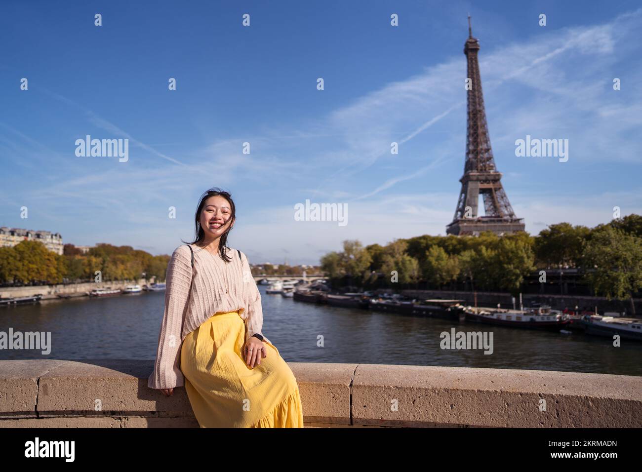 Femme asiatique positive regardant la caméra tout en étant assise sur la frontière de pierre nera la Tour Eiffel en plein soleil Banque D'Images
