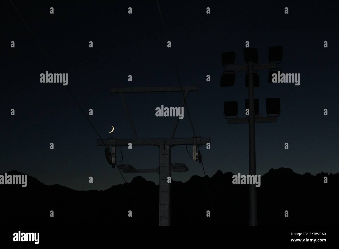 Infrastructure de télécabine avec projecteurs éteints sur la montagne suisse la nuit silhoueté contre le ciel bleu de gradient d'heure avec croissant de lune Banque D'Images