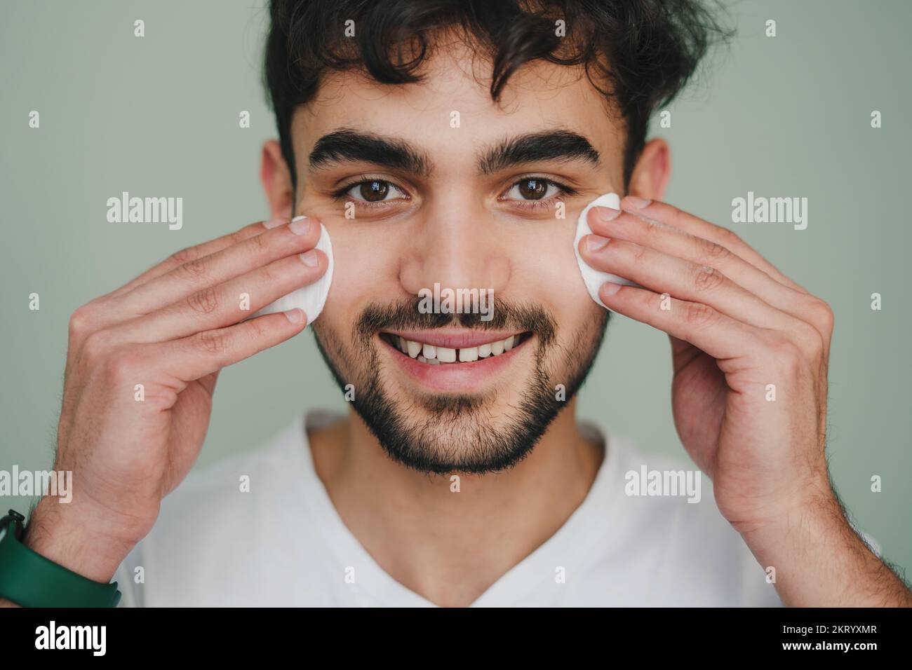 Vue avant portrait d'un homme souriant qui nettoie la peau du visage avec deux tampons de coton regardant l'appareil photo sur fond gris. Portrait isolé Banque D'Images