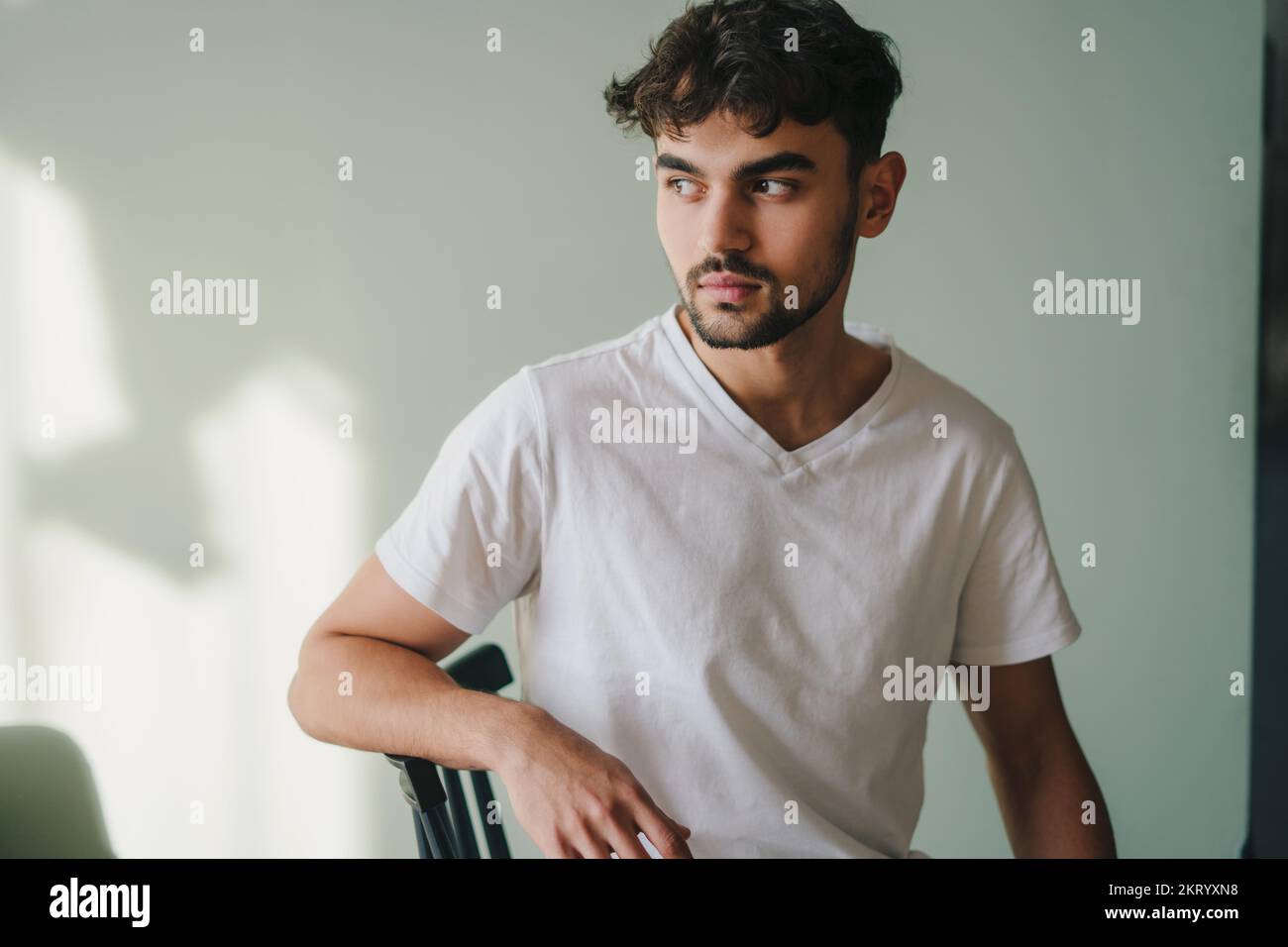 Portrait de beau modèle de mode homme caucasien en t-shirt assis sur une chaise, prise en studio. Personne positive. Appartement confortable. Banque D'Images