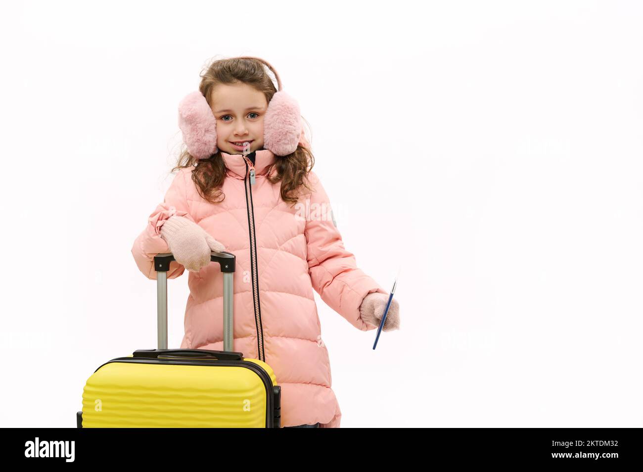 Jolie petite fille portant des couvre-oreilles molletonnés roses, une veste  en duvet et des moufles en laine, avec une valise et une carte  d'embarquement, sur fond blanc Photo Stock - Alamy
