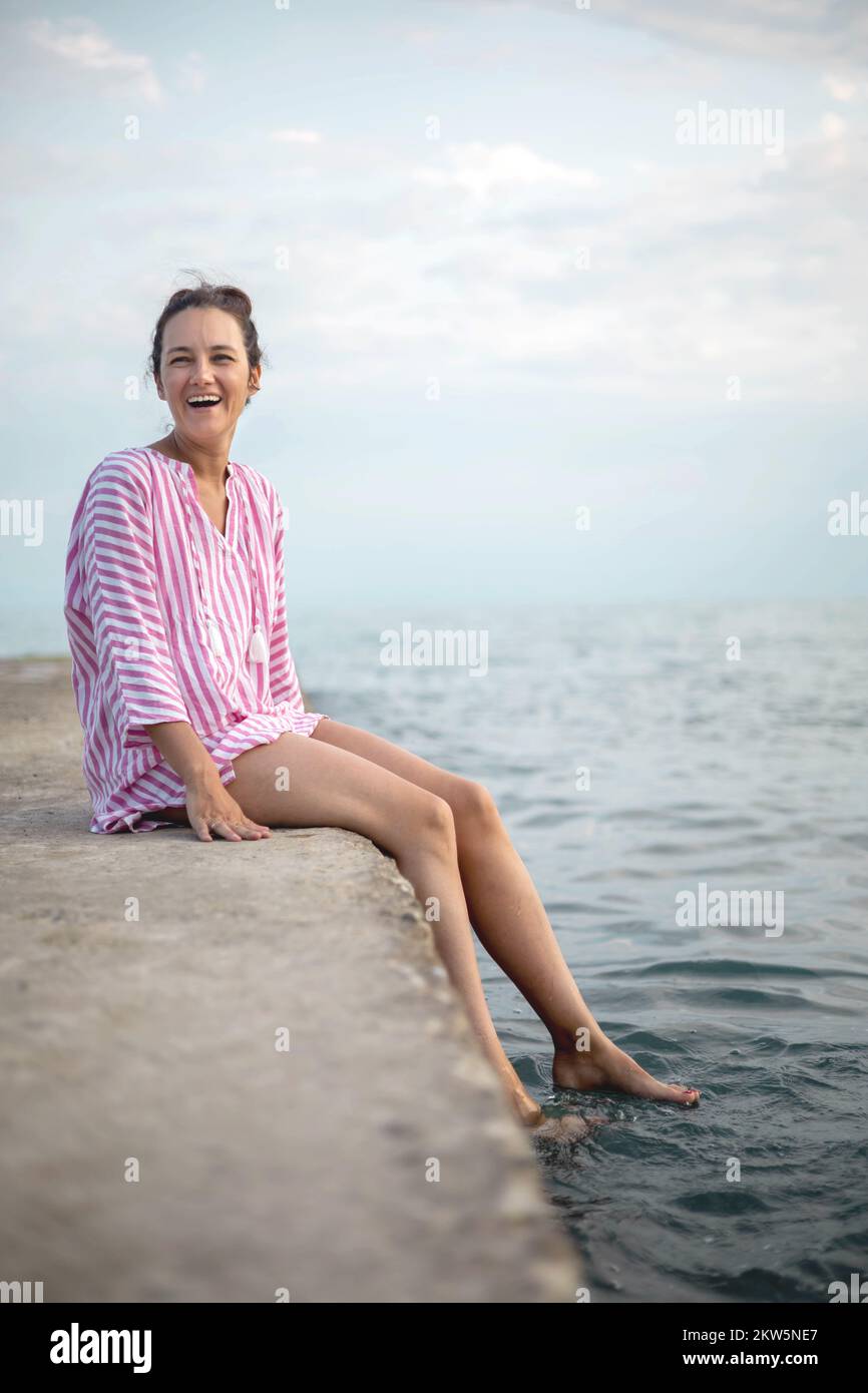 Une jeune femme joyeuse riant assis sur une plage de pierre entourée d'eau éclabousse des vagues mousseuse Banque D'Images