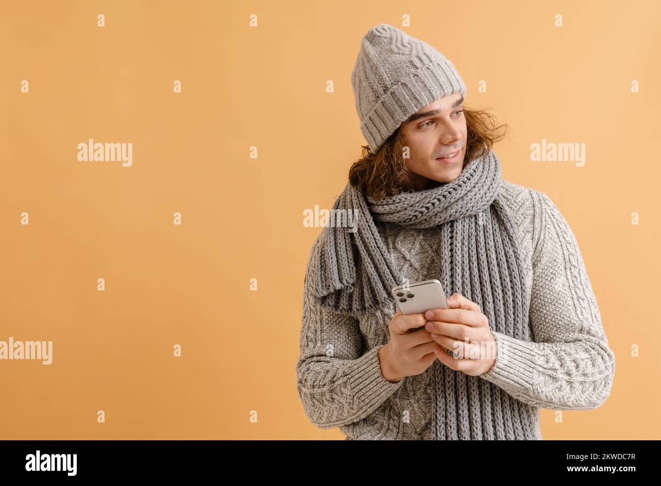 Jeune homme beau avec de longs cheveux en hiver chapeau et écharpe tenant  le téléphone et regardant à droite sur un arrière-plan marron isolé Photo  Stock - Alamy