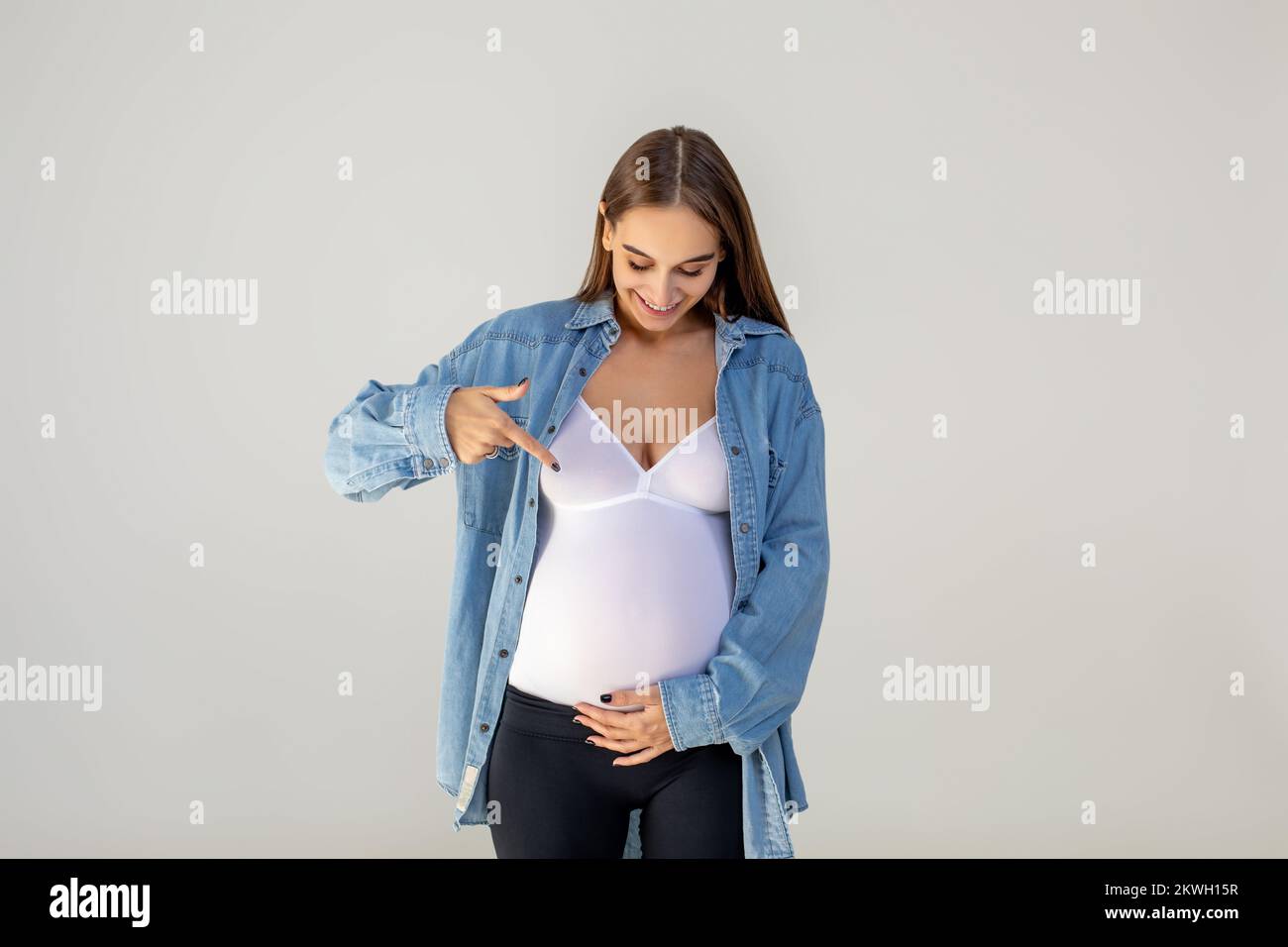 Jeune femme enceinte pointant vers son grand ventre et regardant heureuse Banque D'Images