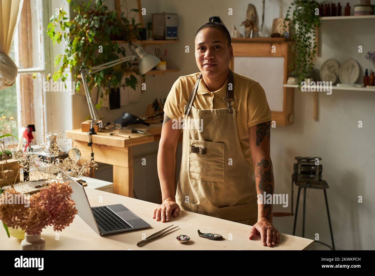 Portrait à la taille haute d'une artiste féminine tatouée regardant l'appareil photo et souriant tout en se tenant dans un studio confortable Banque D'Images