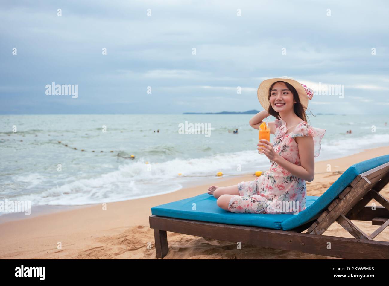 Jeune femme asiatique heureuse portant un chapeau de soleil tenant du jus d'orange se détendant sur une chaise à la plage avec joie. Banque D'Images