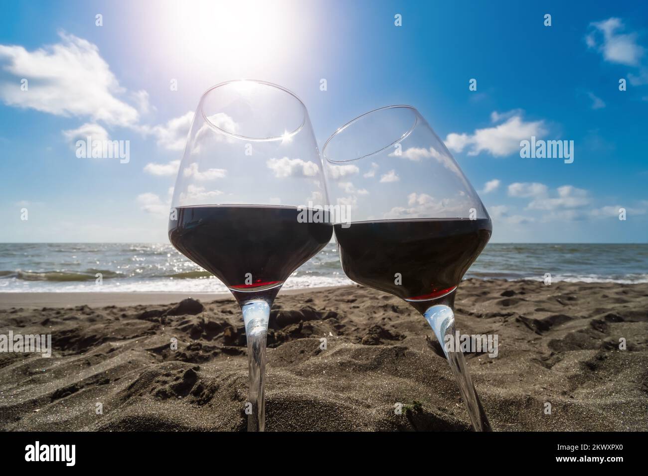 Gros plan de deux verres de vin rouge se tenant dans le sable de la plage contre un ciel bleu avec des nuages et des rayons du soleil Banque D'Images