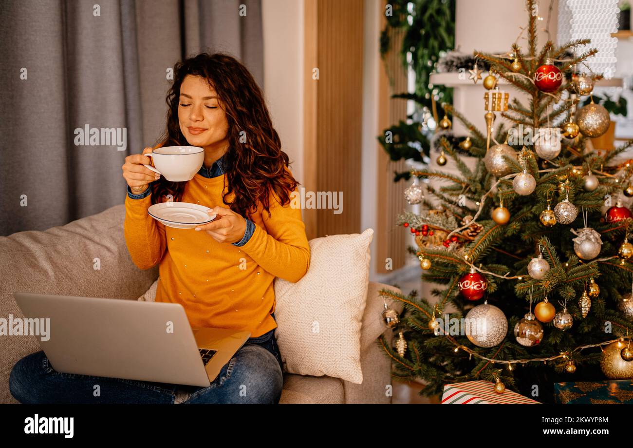 Jeune femme célébrant seul Noël tranquille, utilisant un ordinateur portable, regardant un film de Noël, buvant du café près des boîtes-cadeaux à la maison. Vacances d'hiver, nouvel an e Banque D'Images