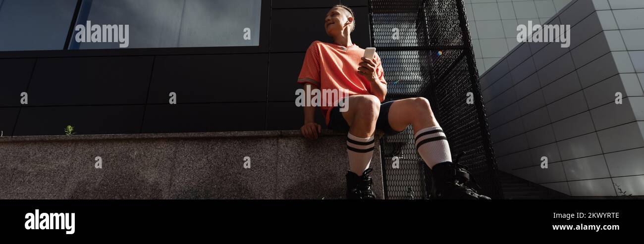 Vue à angle bas d'un jeune homme dans des rouleaux tenant un smartphone et écoutant de la musique dans un écouteur dans une rue urbaine, bannière, image de stock Banque D'Images