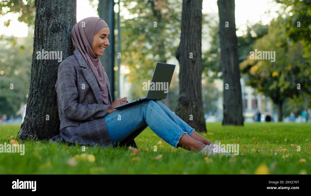 Bonne femme d'affaires musulmane islamique dans le hijab assis près de l'arbre sur l'herbe verte dans le parc de la ville discutant avec des amis de la famille à distance conversation Banque D'Images