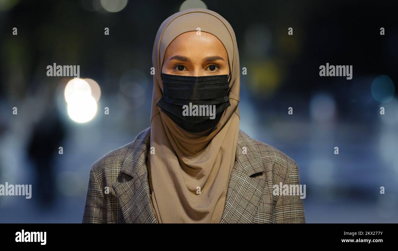 Une femelle islamique en gros plan dans un masque médical posant dans une ville nocturne. Femme d'affaires musulmane portant le hijab fille masquée debout regardant la caméra soir urbaine Banque D'Images