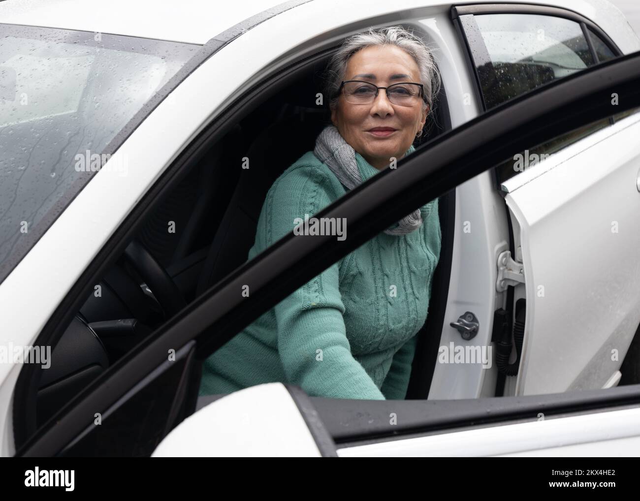 Une dame aux cheveux gris s'assoit sur le siège du conducteur dans une voiture avec une porte ouverte et sourit à la caméra Banque D'Images