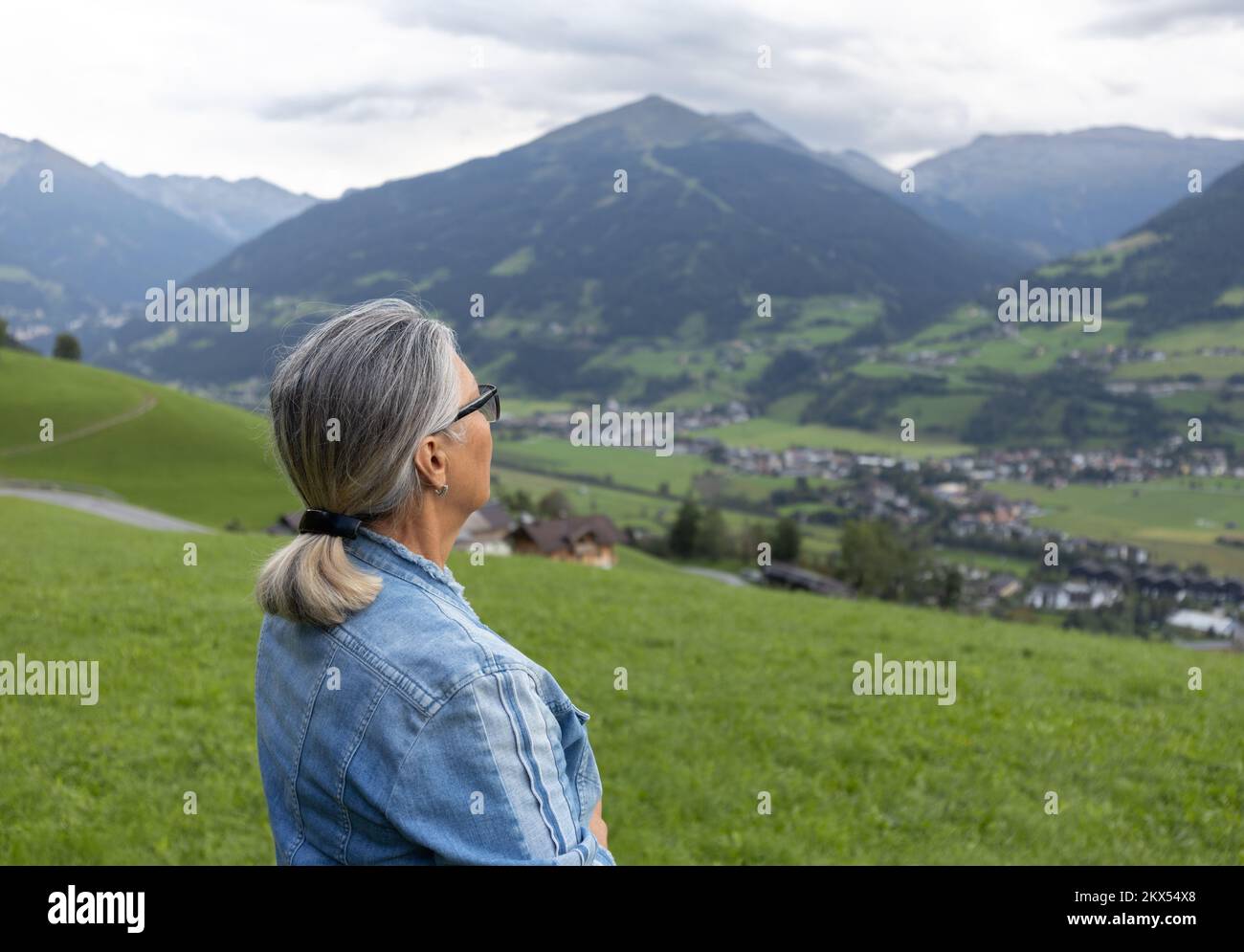 Une femme aux cheveux gris en costume denim se tient près de la route sur une pente herbeuse et regarde vers les montagnes. Banque D'Images