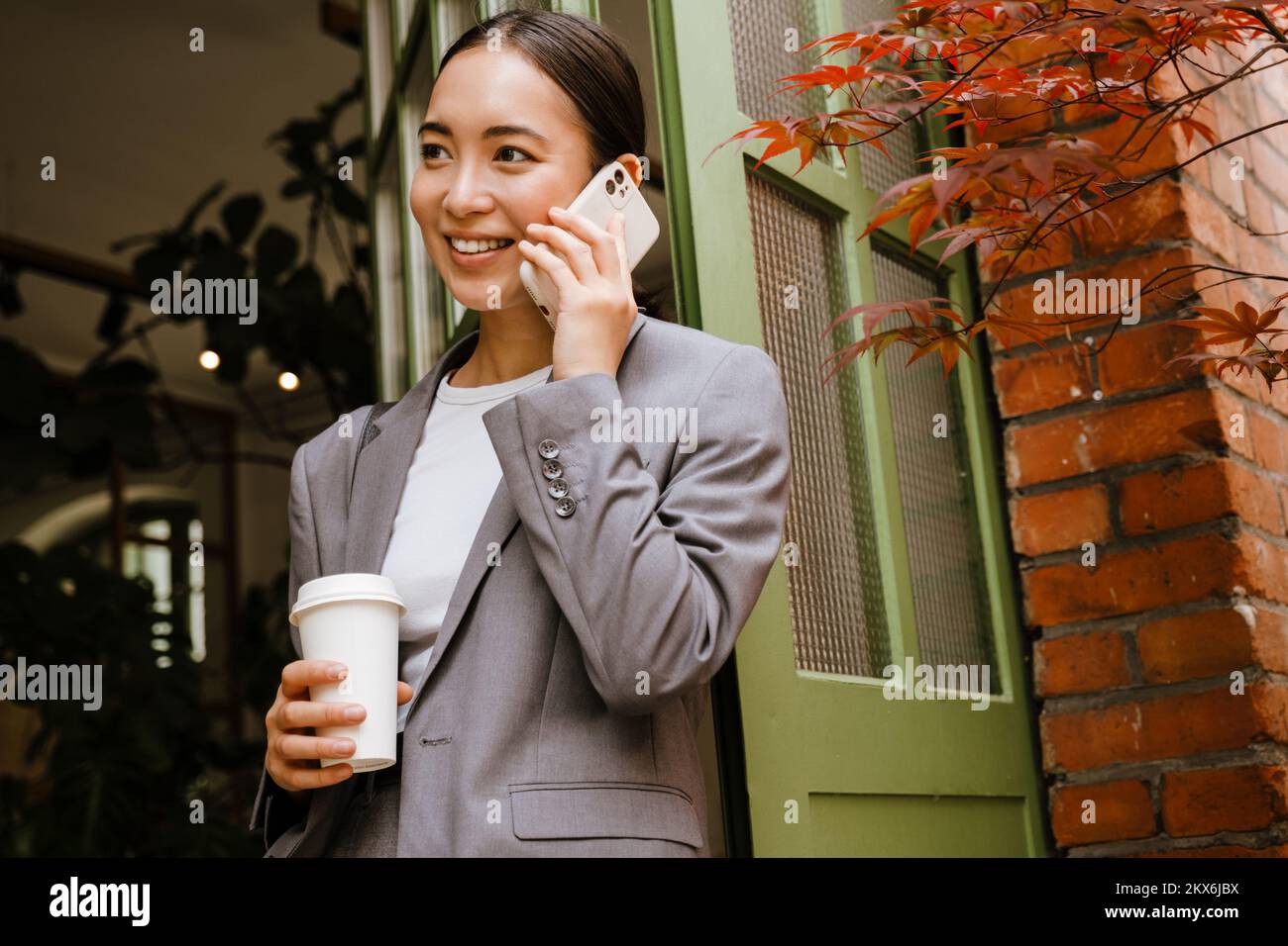 Jeune femme d'affaires asiatique souriant et parlant au téléphone portable tout en se tenant près de la porte dans la rue Banque D'Images