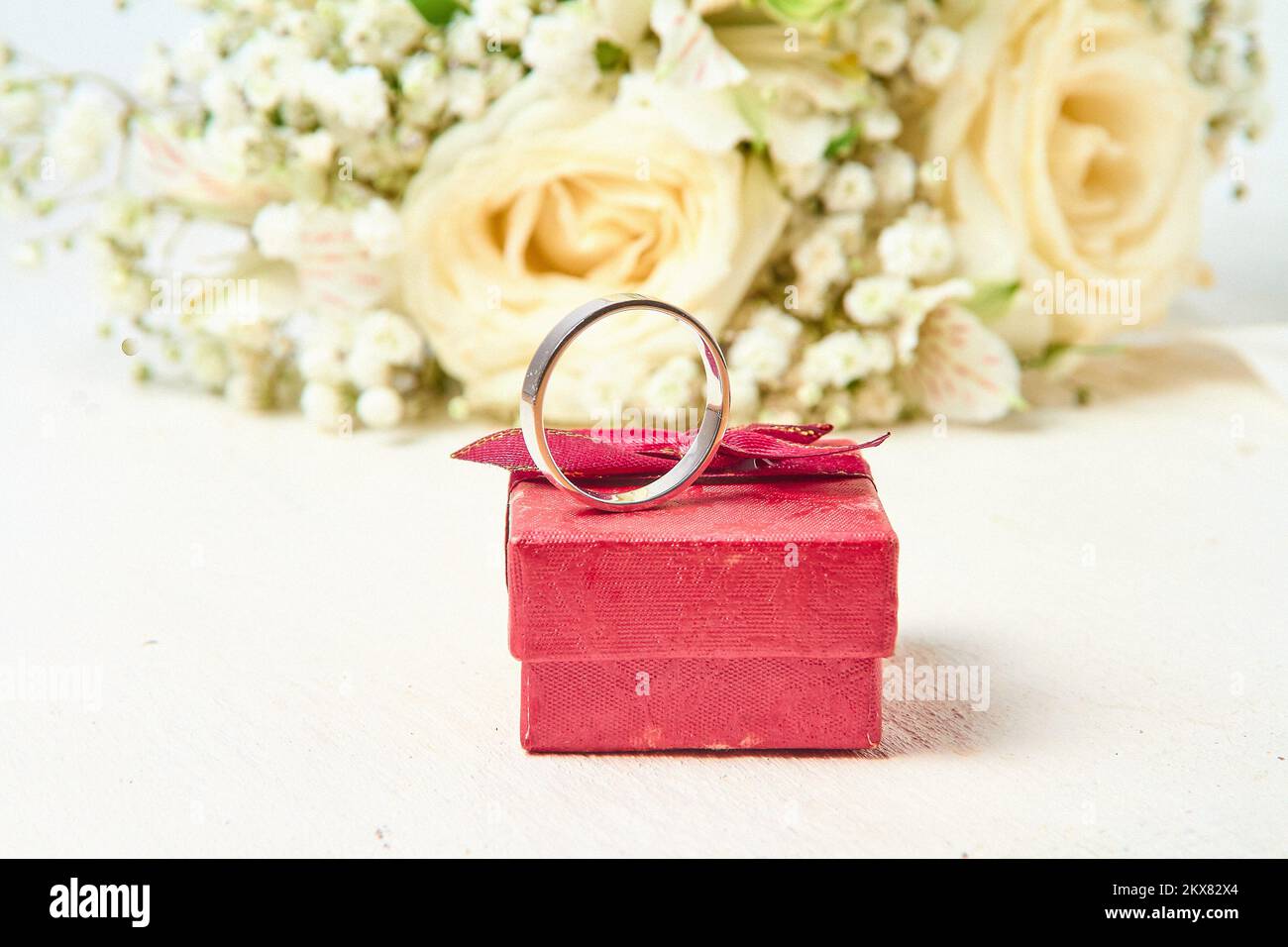 Un anneau d'or blanc se dresse sur une boîte cadeau rouge et un bouquet de fleurs sur un fond d'arbre blanc texturé, vacances. Banque D'Images