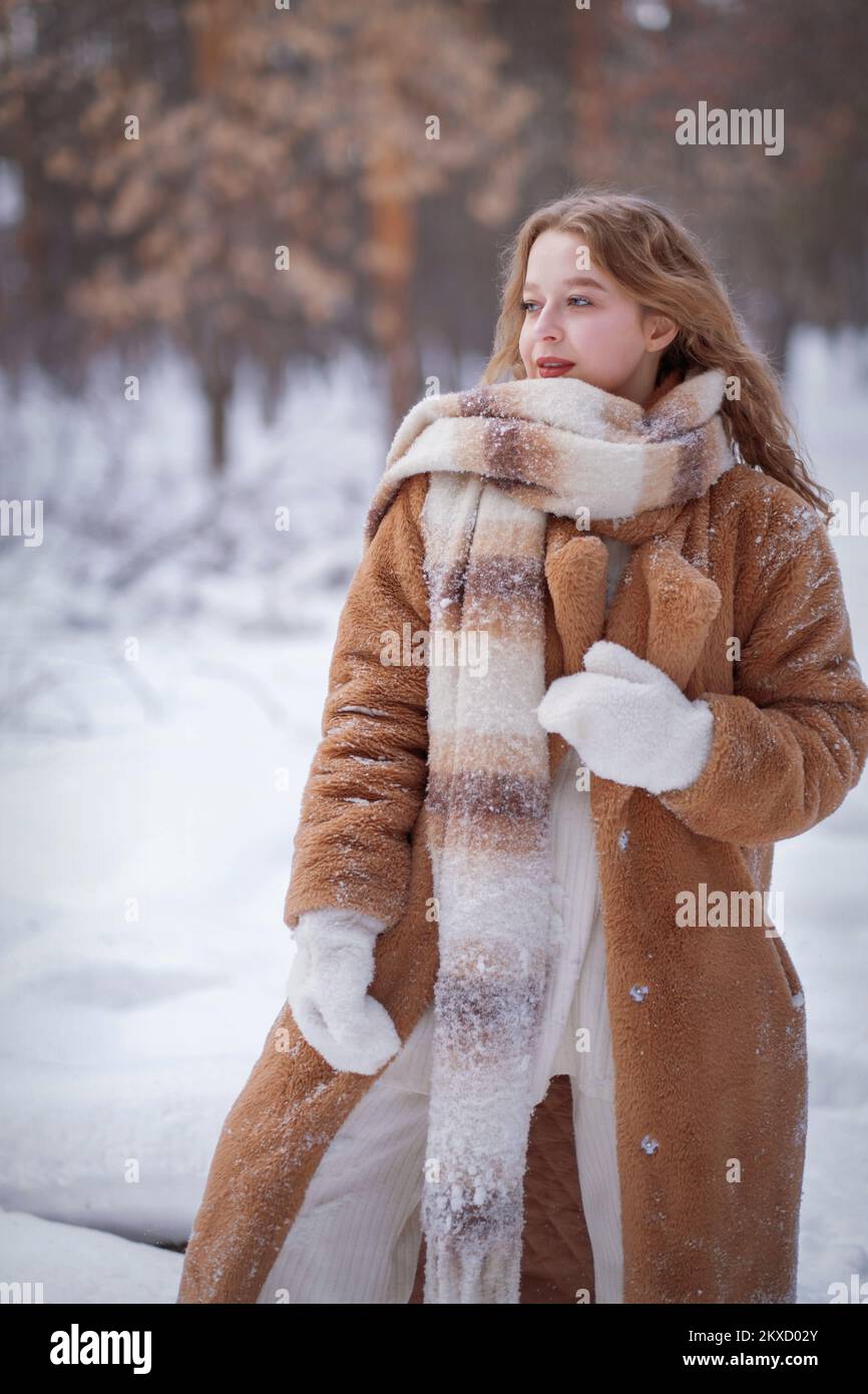 Une blonde blanche en bonnet tricoté, une longue écharpe et un manteau de  fourrure se promène dans la forêt et sourit. Femme attrape la neige dans  des moufles blanches. L'hiver est dans