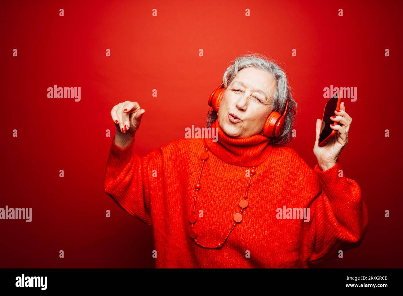 Femme âgée vêtue de vêtements rouges, avec casque rouge et smartphone rouge, dansant sur fond rouge Banque D'Images Femme âgée vêtue de vêtements rouges, avec casque rouge et smartphone rouge, dansant sur fond rouge Banque D'Images