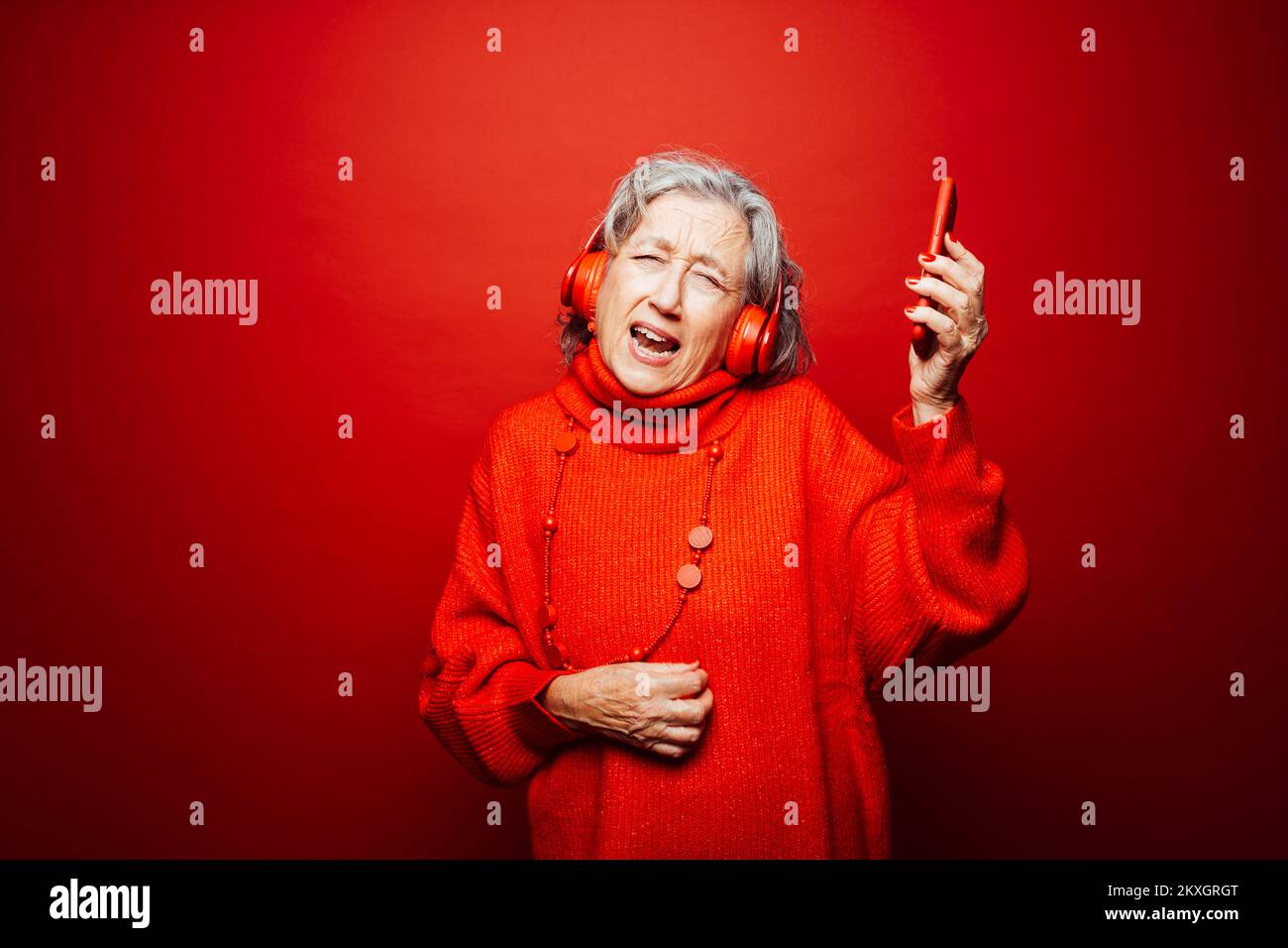 Femme âgée vêtue de vêtements rouges, avec casque rouge et smartphone rouge, dansant sur fond rouge Banque D'Images Femme âgée vêtue de vêtements rouges, avec casque rouge et smartphone rouge, dansant sur fond rouge Banque D'Images