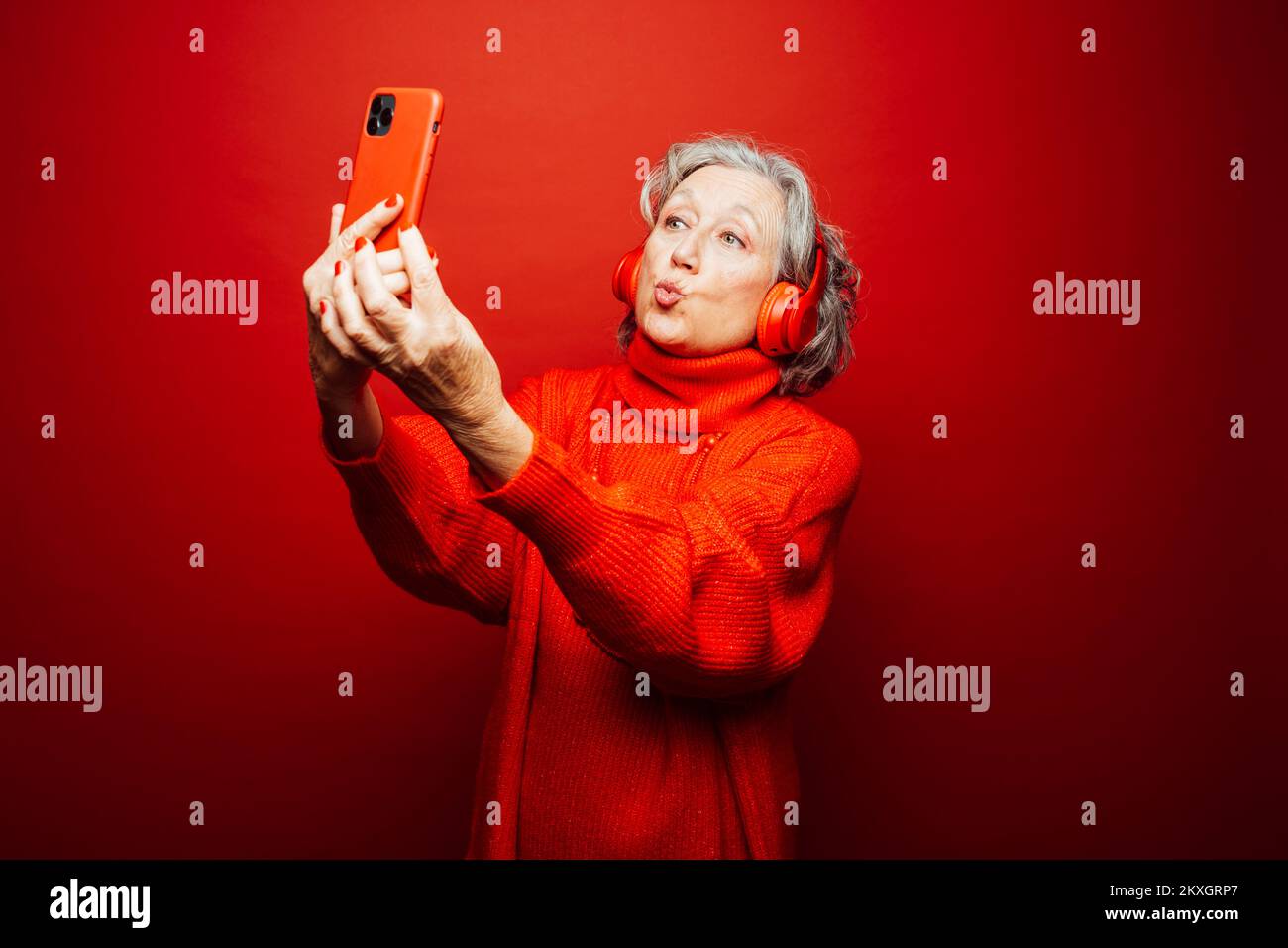 Femme âgée vêtue de vêtements rouges, emportant un selfie avec un casque rouge et un smartphone rouge, sur un fond rouge Banque D'Images Femme âgée vêtue de vêtements rouges, emportant un selfie avec un casque rouge et un smartphone rouge, sur un fond rouge Banque D'Images