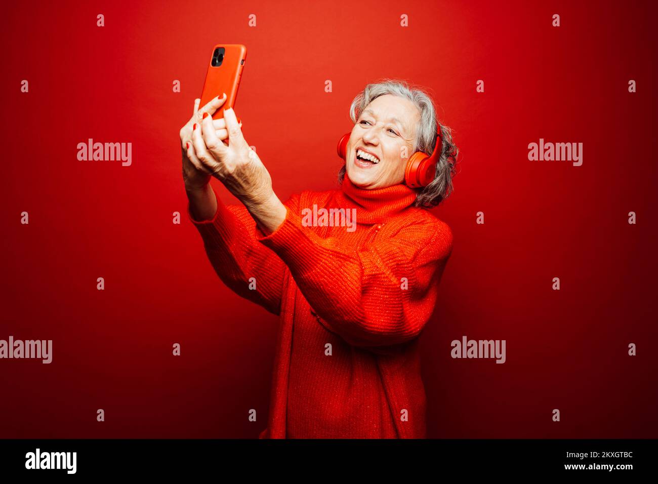Femme âgée vêtue de vêtements rouges, emportant un selfie avec un casque rouge et un smartphone rouge, sur un fond rouge Banque D'Images