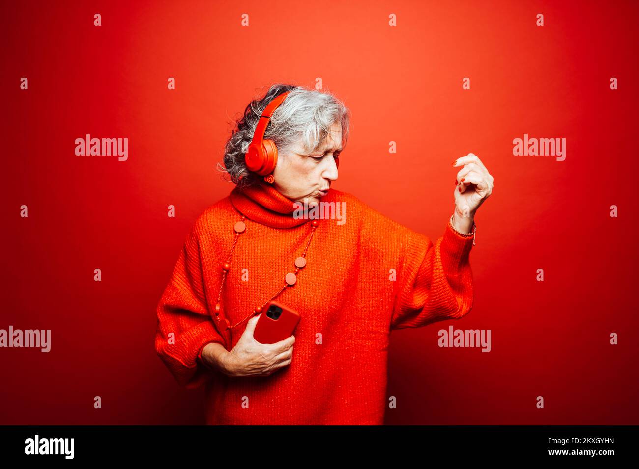 Femme âgée vêtue de vêtements rouges, avec casque rouge et smartphone rouge, dansant sur fond rouge Banque D'Images Femme âgée vêtue de vêtements rouges, avec casque rouge et smartphone rouge, dansant sur fond rouge Banque D'Images
