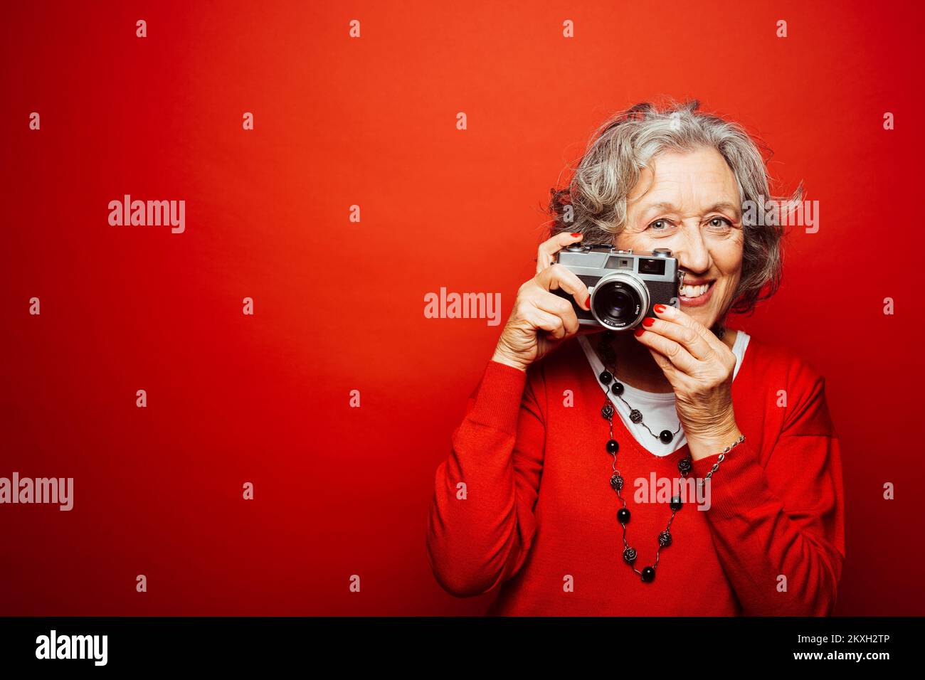 Femme âgée vêtue de vêtements rouges, prenant des photos avec un ancien appareil photo, sur fond rouge Banque D'Images