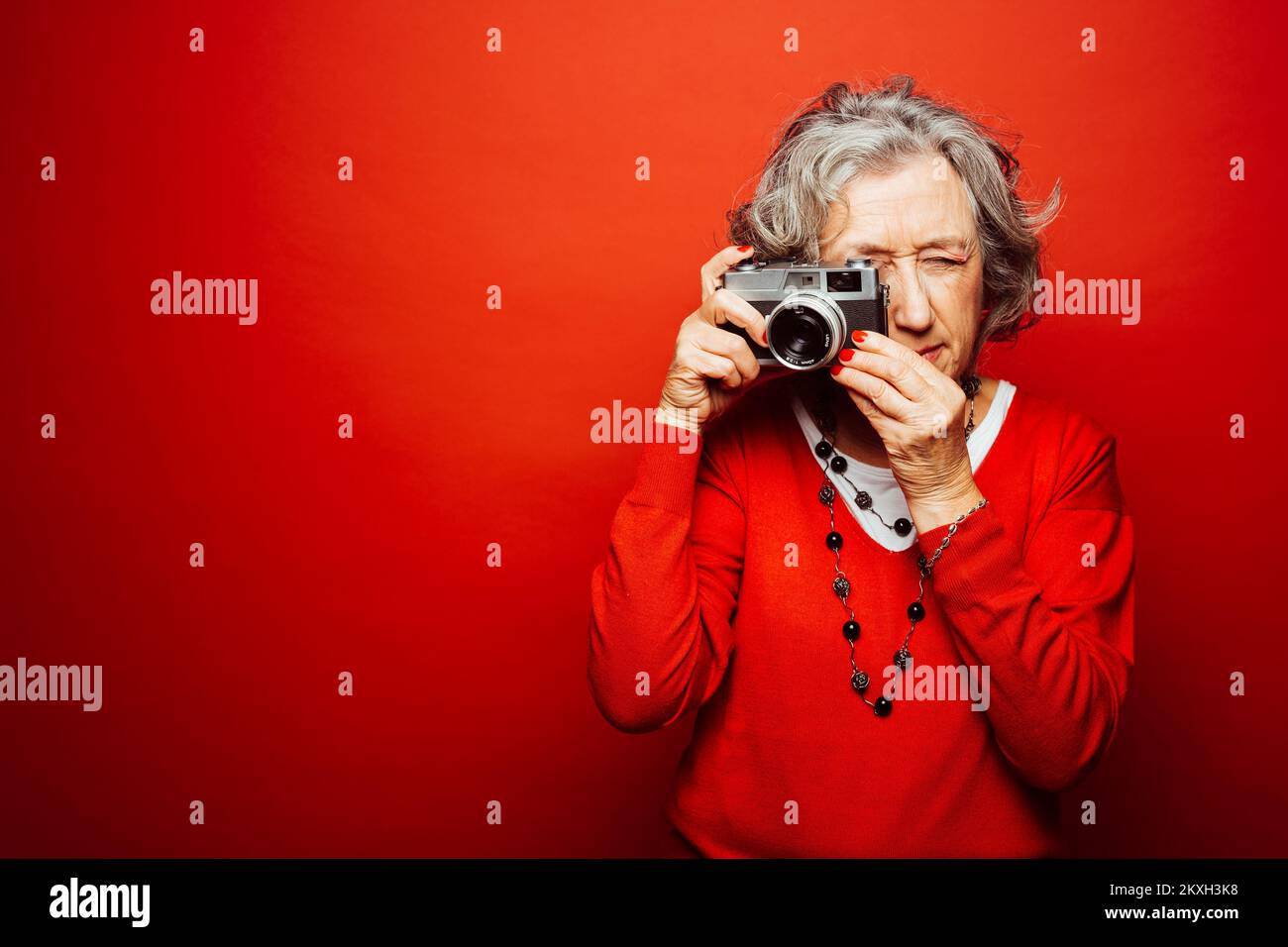 Femme âgée vêtue de vêtements rouges, prenant des photos avec un ancien appareil photo, sur fond rouge Banque D'Images Femme âgée vêtue de vêtements rouges, prenant des photos avec un ancien appareil photo, sur fond rouge Banque D'Images