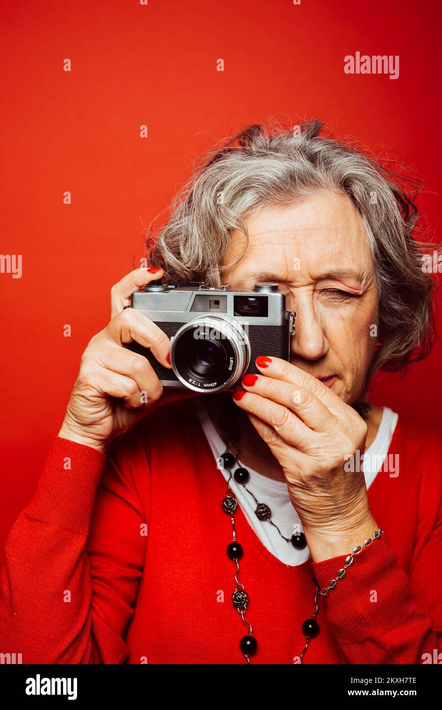 Femme âgée vêtue de vêtements rouges, prenant des photos avec un ancien appareil photo, sur fond rouge Banque D'Images Femme âgée vêtue de vêtements rouges, prenant des photos avec un ancien appareil photo, sur fond rouge Banque D'Images