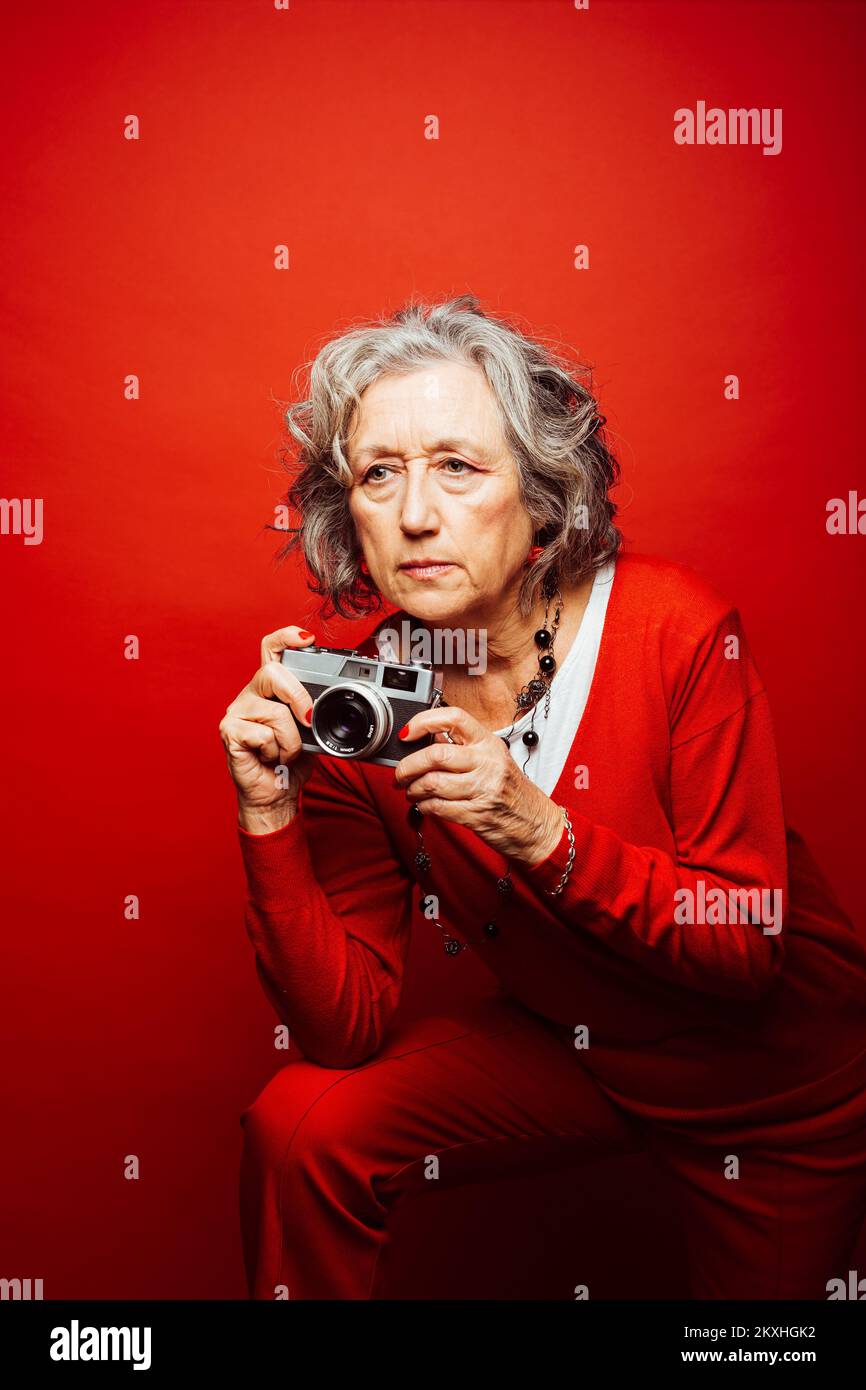 Femme âgée vêtue de vêtements rouges, prenant des photos avec un ancien appareil photo, sur fond rouge Banque D'Images Femme âgée vêtue de vêtements rouges, prenant des photos avec un ancien appareil photo, sur fond rouge Banque D'Images
